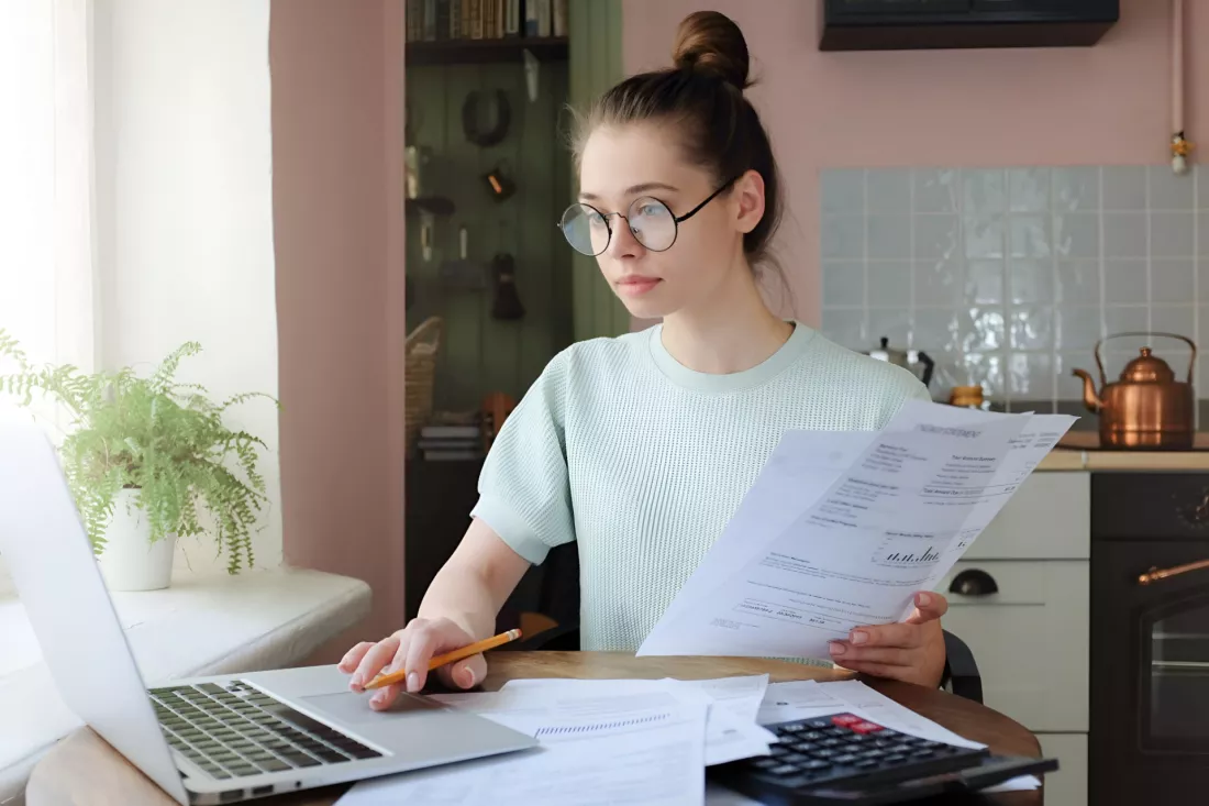 Mloda kobieta w okularach przeglada dokumenty przy laptopie i kalkulatorze w kuchni